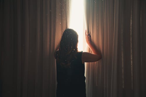 Free Back view of anonymous lady opening ribbed curtain behind window with glowing light while standing in apartment Stock Photo