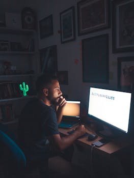 A man deep in thought working at a computer in a dimly lit home office setting.