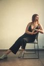 Young contemplative tattooed female with big breasts in stylish wear and sneakers sitting on chair in flat and looking away