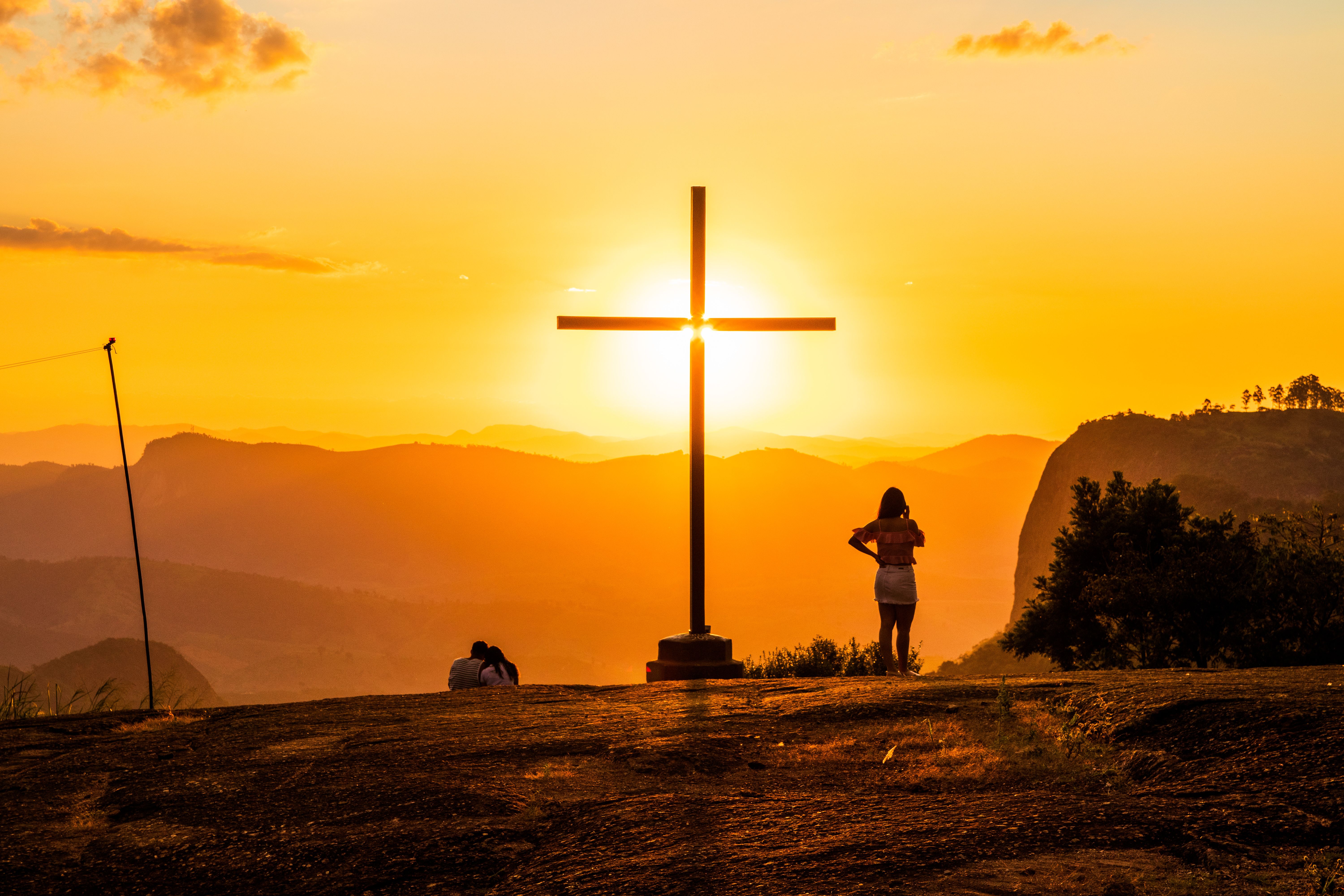 Back view of anonymous travelers standing near cross behind mountains under colorful sky with shining sun at sunset — Foto: Marcos Luis  Leonardo / Pexels