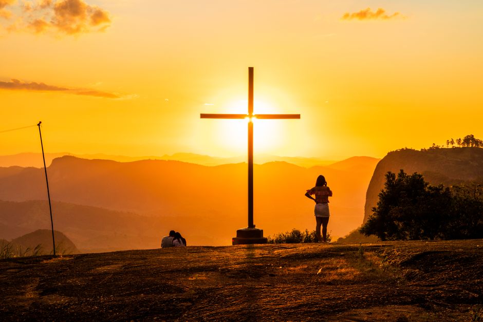 Back view of anonymous travelers standing near cross behind mountains under colorful sky with shining sun at sunset — Foto: Marcos Luis Leonardo / Pexels