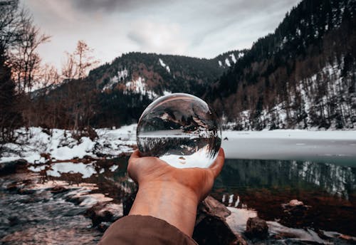 Crop man with crystal ball near lake in snowy mountains