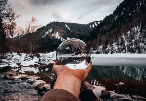 Crop unrecognizable male holding transparent glass ball while standing near pond reflecting mounts under cloudy sky in winter
