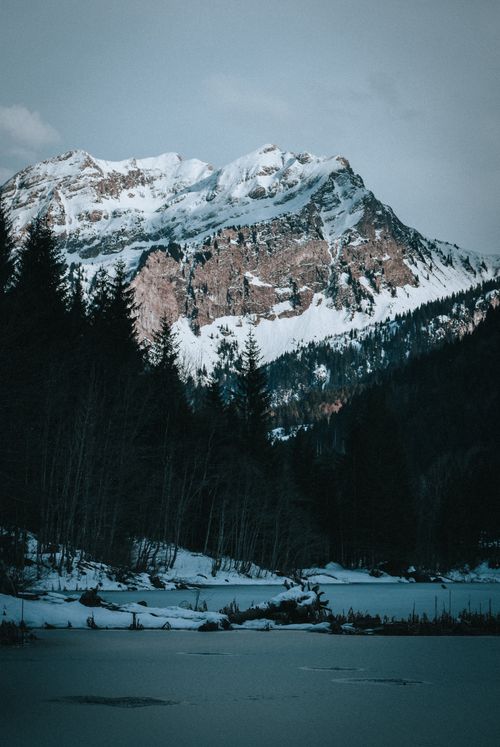 Free Picturesque view of high mountains with snow near growing trees and frozen lake under cloudy sky in wintertime Stock Photo
