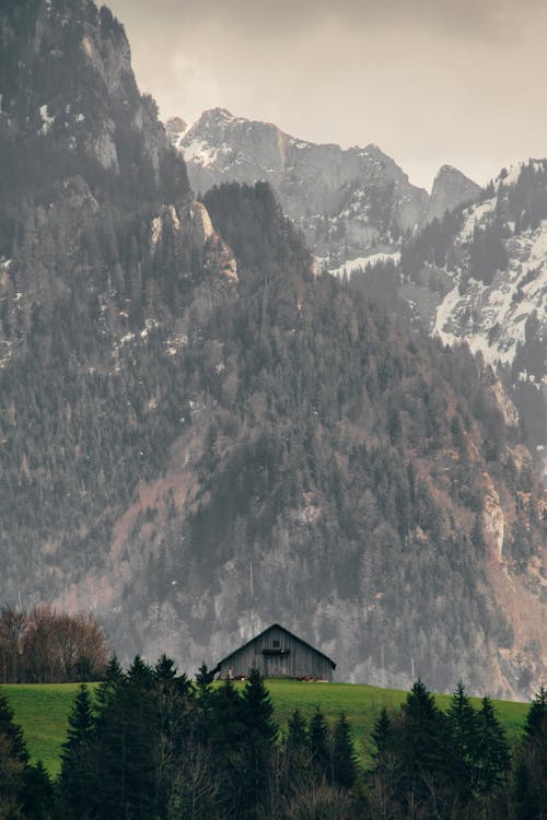 Spectacular view of snowy ridge near bright green lawn with aged building and growing trees under cloudy sky