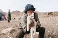 Smiling Mongolian boy in casual clothes wearing warm hat and and showing local white cat and looking at camera near yurt