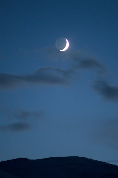 Bright thin crescent moon hanging over hilly countryside in dark cloudy sky at sunset