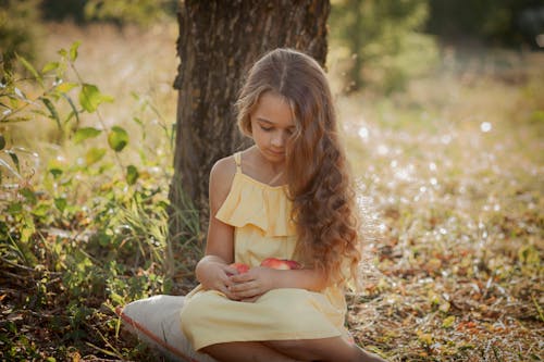 Thoughtful little girl in casual dress sitting on grass near tree trunk in sunny day and looking down