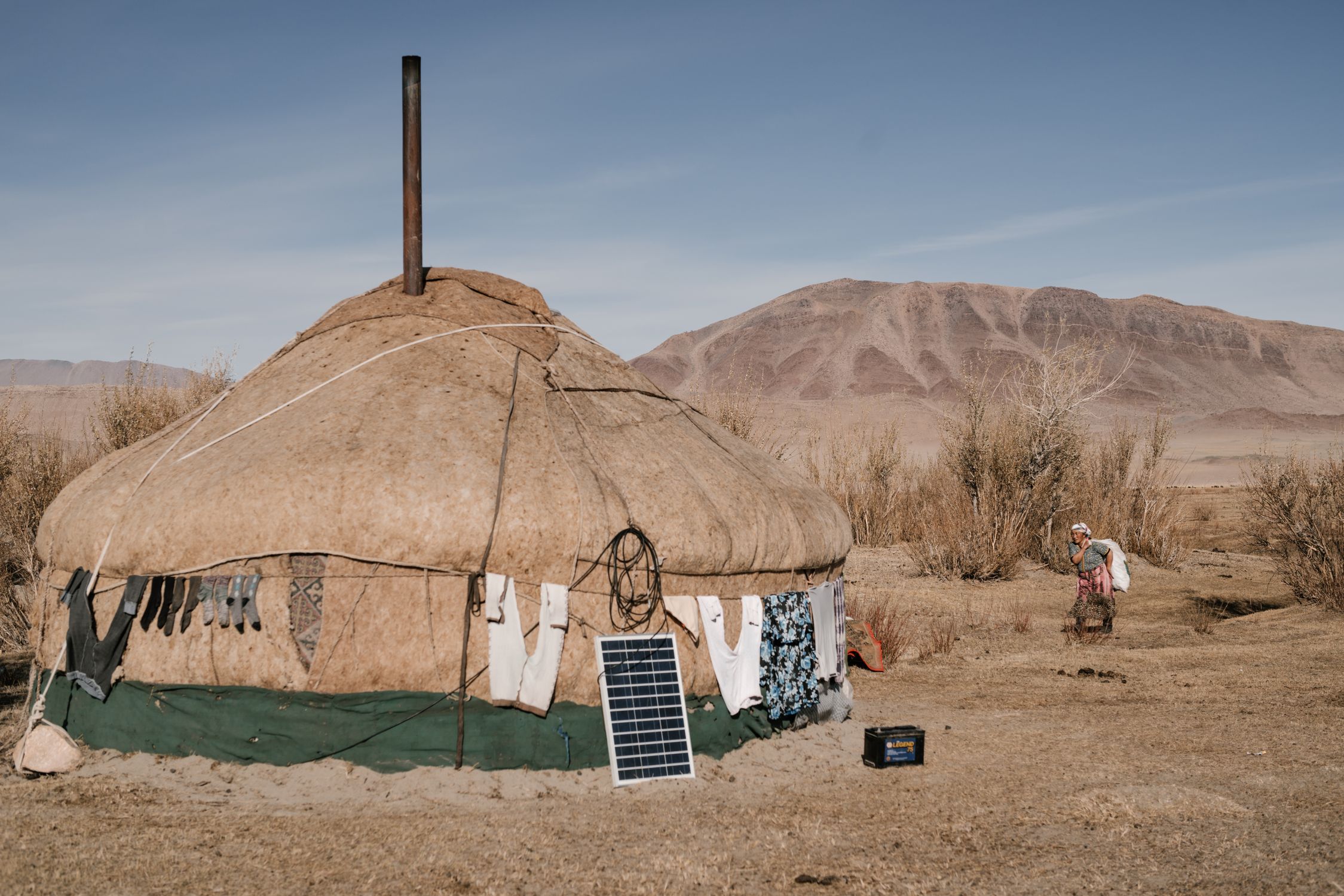 Exterior of traditional Mongolian yurt with linen hanging on ropes around and woman walking nearby in sunlght