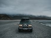 SUV automobile with headlights on parked on sand beach near ocean and ridge covered with mist in evening time