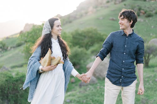 Young positive woman with loaves of bread in romantic dress and kerchief holding hand of boyfriend and looking at each other while walking in grassy rural field