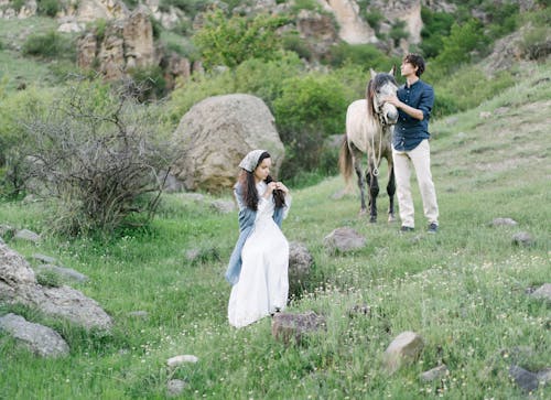 Full length of young romantic couple in casual clothes relaxing on grassy hillside after horse ride in summer day