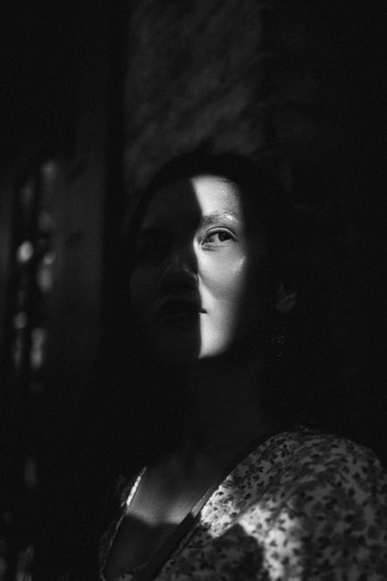 Black and white contemplative calm Asian female in casual clothes standing with shadows on face near brick building and looking away suspiciously