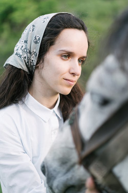 Young pensive female with long dark hair in kerchief standing in paddock and taking care of horse in summer day