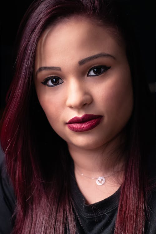 Crop calm Asian female with dyed hair wearing bright makeup and black shirt looking at camera while in dark room