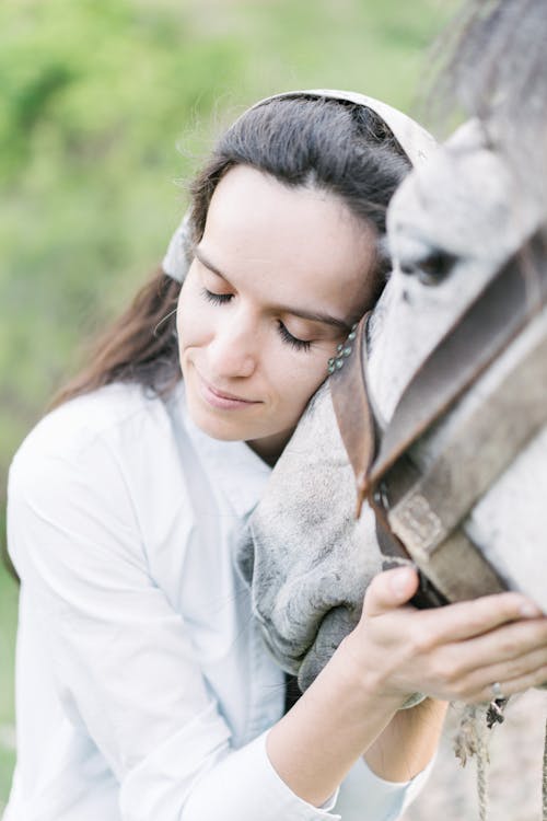 Cheerful young female in white shirt smiling with closed eyes while caressing obedient horse on pasture