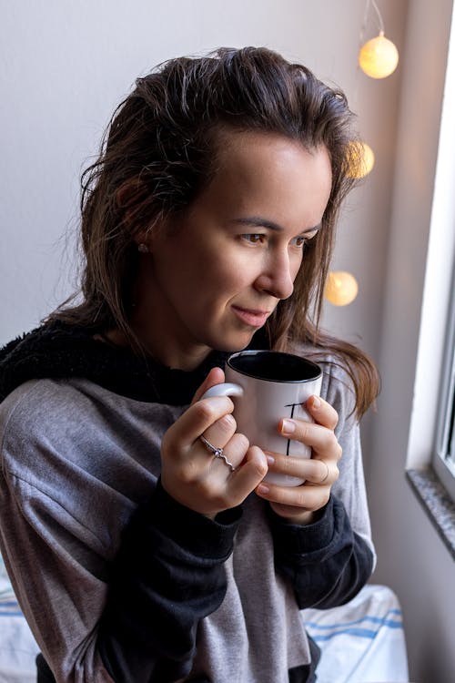 Free Positive smiling female in casual comfy wear drinking hot aromatic beverage and looking out window Stock Photo