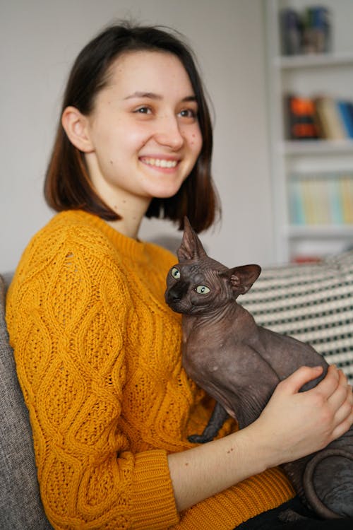 Side view of positive young female in warm sweater relaxing on sofa with Sphynx cat in hands and looking away