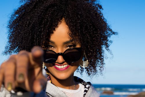 Crop happy young African American female with bright makeup and curly hair in stylish sunglasses standing on seashore and pointing finger at camera