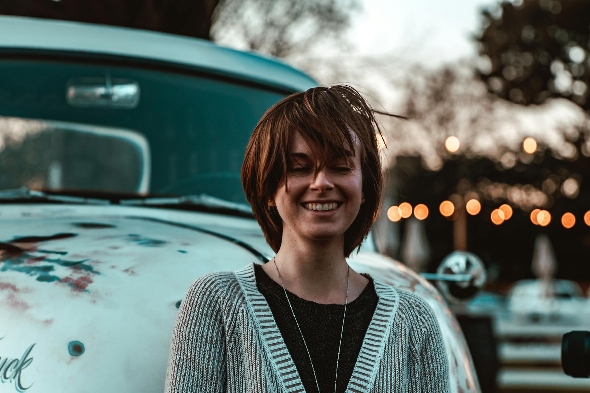 Optimistic smiling female in casual comfy outfit standing with eyes closed against old fashioned truck in evening