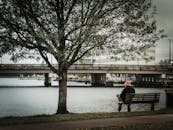 一位男性背对镜头，身穿保暖外套、头戴帽子，双腿交叉坐在木凳上，在阴沉的秋日里眺望城市河景