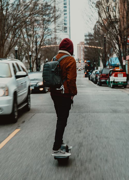 Back view full length anonymous male skater in warm casual wear and hood riding skateboard along asphalt road in urban environment