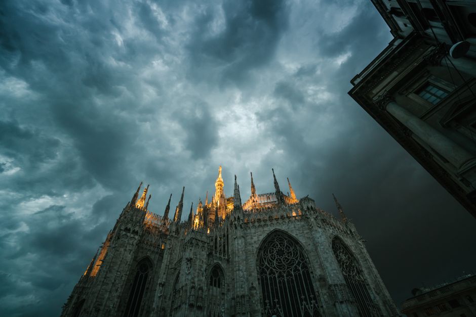 Low angle of Metropolitan Cathedral Basilica of Nativity of Saint Mary in Milan with sharp spires lit up with bright light against gloomy overcast sky