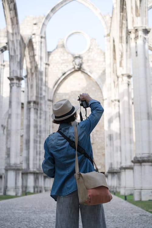 Free Back view of anonymous female tourist in hat taking photo of pointed arch of Convent of Lady of Mount Carmel in Lisbon on sunny day Stock Photo