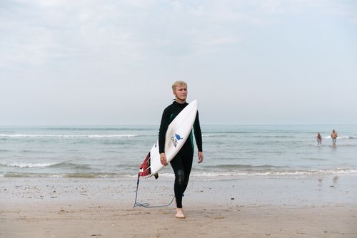 Free Confident male surfer in black wetsuit walking on wet ocean shore carrying surfboard under arm on cloudy summer day Stock Photo