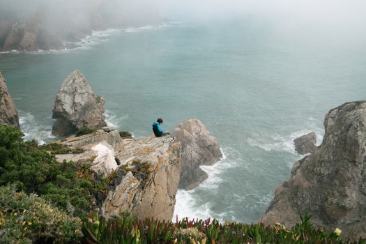 From above of anonymous male traveler sitting on rocky edge of cliff above sea