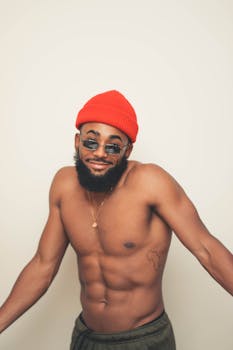 A confident young man poses shirtless with a red hat and sunglasses, showcasing a cool, casual style.