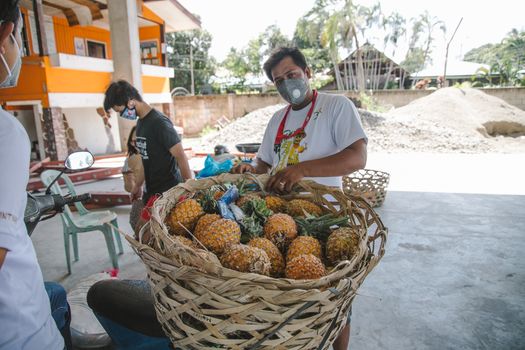 A man wearing a mask sells fresh pineapples from a basket outdoors.