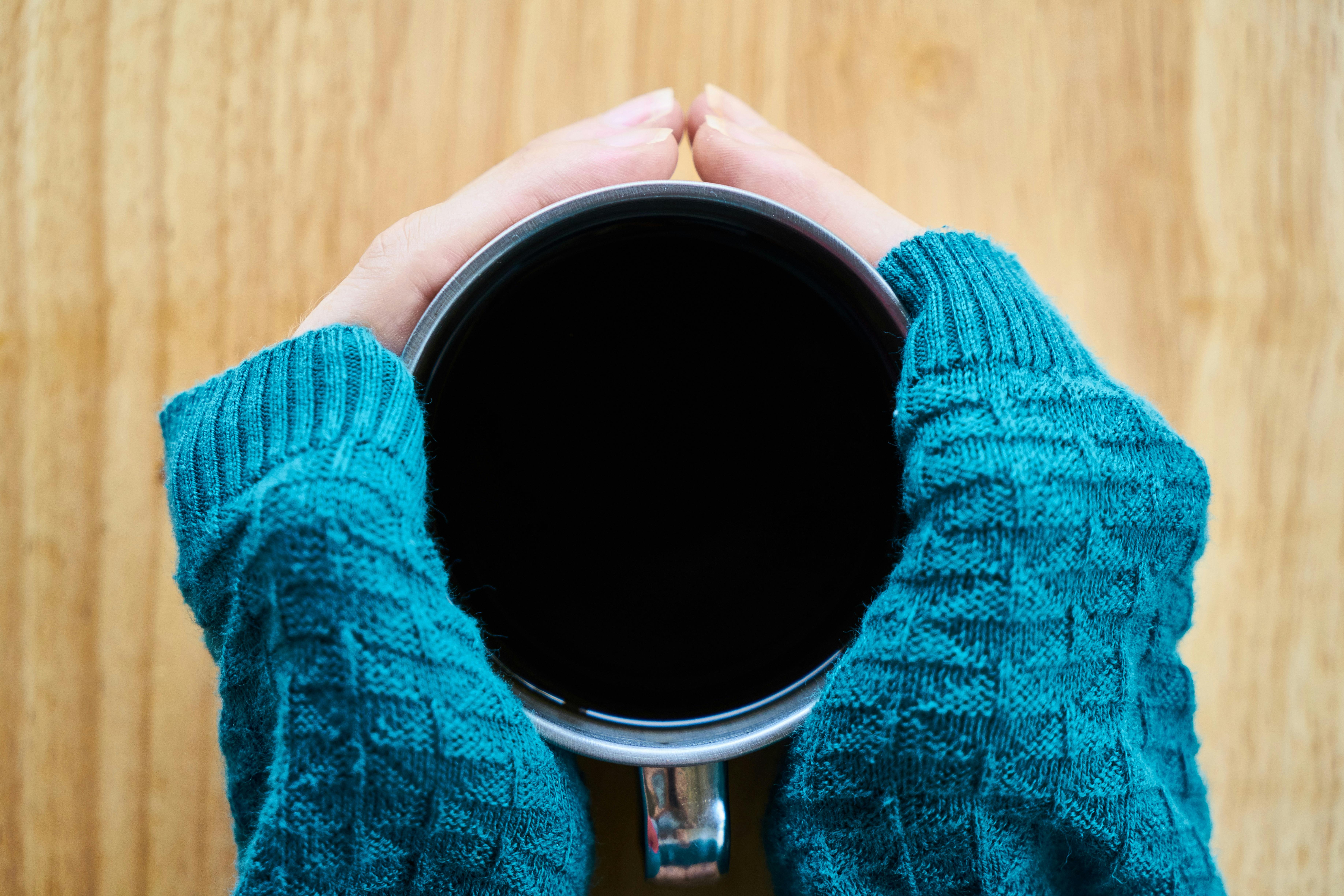 Close-up of hands holding a coffee cup, wrapped in a cozy teal sweater, perfect for a warm comfort theme.