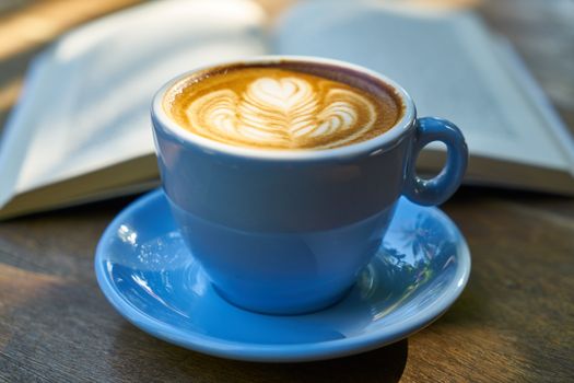 Close-up of a latte in a blue cup with beautiful art, next to an open book on a wooden table at a café.