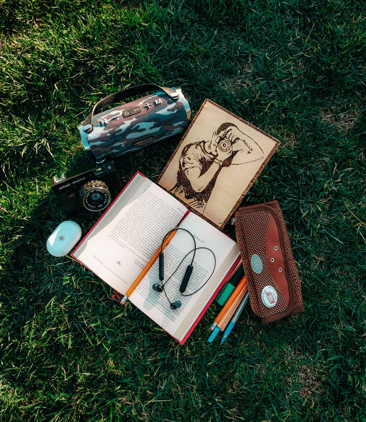 Opened Book And Painting On Green Grass With Wireless Speaker And Photo Camera