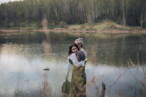Free Side view of gentle romantic couple embracing each other while standing near pond with reflecting water Stock Photo