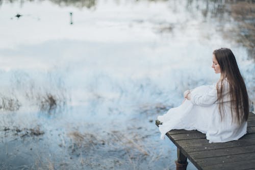 Side view of peaceful young female wearing elegant dress sitting on wooden boardwalk near lake