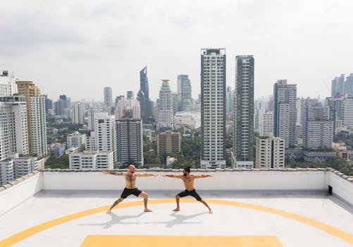 Two men practice yoga poses on a rooftop with a city skyline in the background.