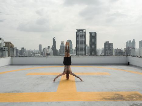 Shirtless man practicing a headstand on a rooftop helipad with a city skyline background, promoting wellness and balance.