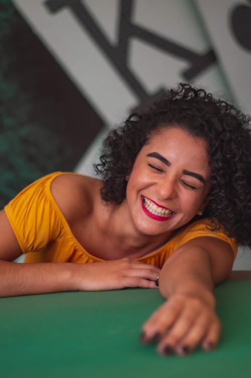 Young ethnic lady with red lips laughing with eyes closed while leaning on green table