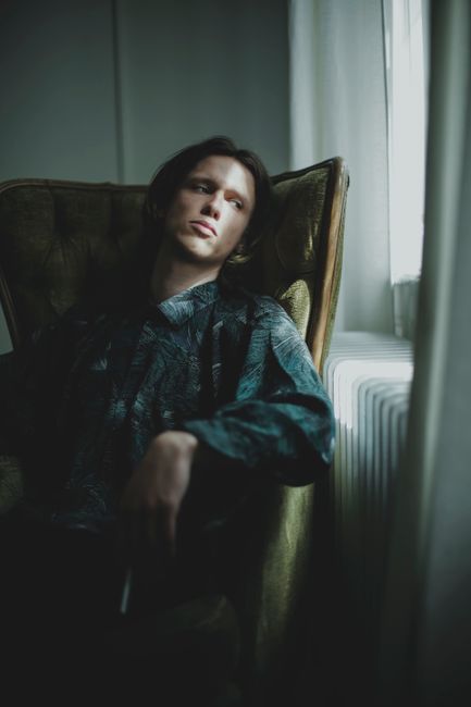Lonely young guy relaxing in comfortable armchair and looking out window in dark room at home