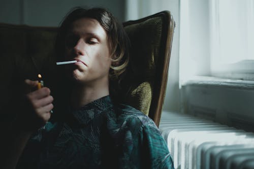 Young guy lighting cigarette and smoking while sitting in armchair near window at home
