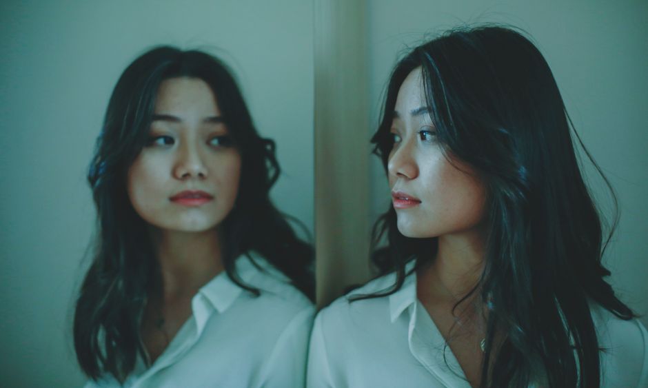 Pretty young Asian woman leaning on wall and looking at reflection in mirror