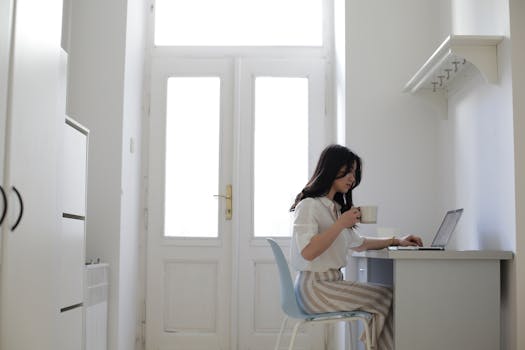 Asian woman working remotely on a laptop at a home desk with a cup of coffee. Bright and cozy workspace.