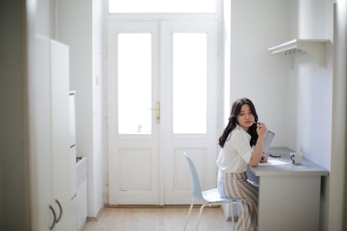 Side view of young Asian woman in casual clothes biting pen and thinking while sitting at table and doing homework assignment at home
