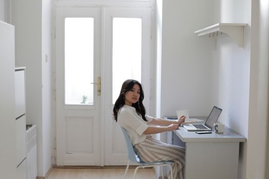 Side view of young Asian woman in casual clothes sitting at desk with laptop and stationery and looking at camera while working on remote project at home