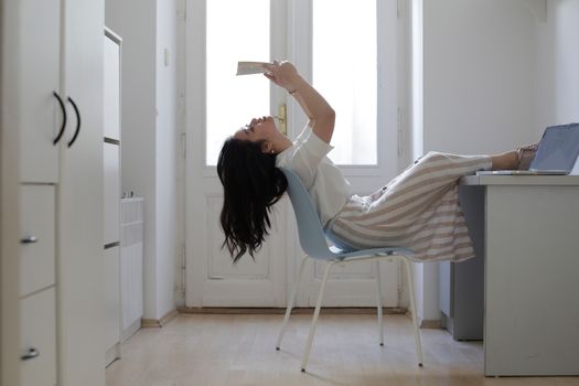 Side view of young Asian woman in casual clothes leaning back on chair and reading document while doing homework at home