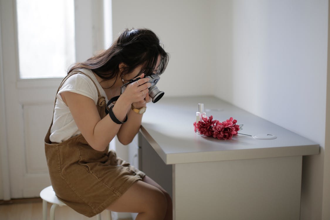 Side view of young Asian female shooting bunch of fresh flowers and bottle of perfume on table at home