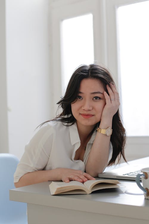 Free Young Asian woman smiling and looking at camera while sitting at desk and reading book during studies at home Stock Photo
