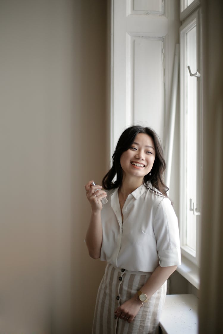 Cheerful Asian Female Applying Perfume Near Window